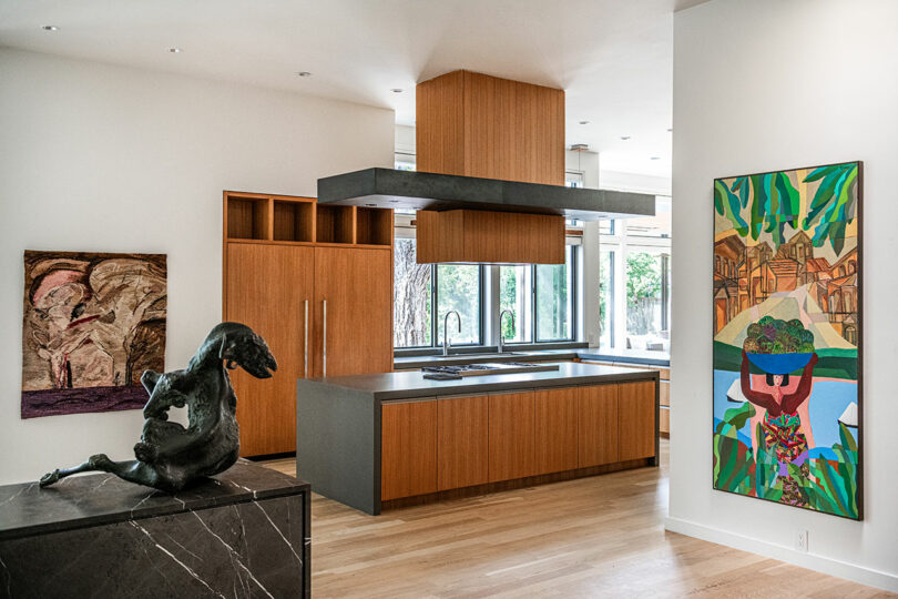 Modern kitchen with wooden cabinets, a kitchen island, and large artworks on the walls, reflecting Brazilian Modernism. A black sculpture is placed on a dark countertop in the foreground.
