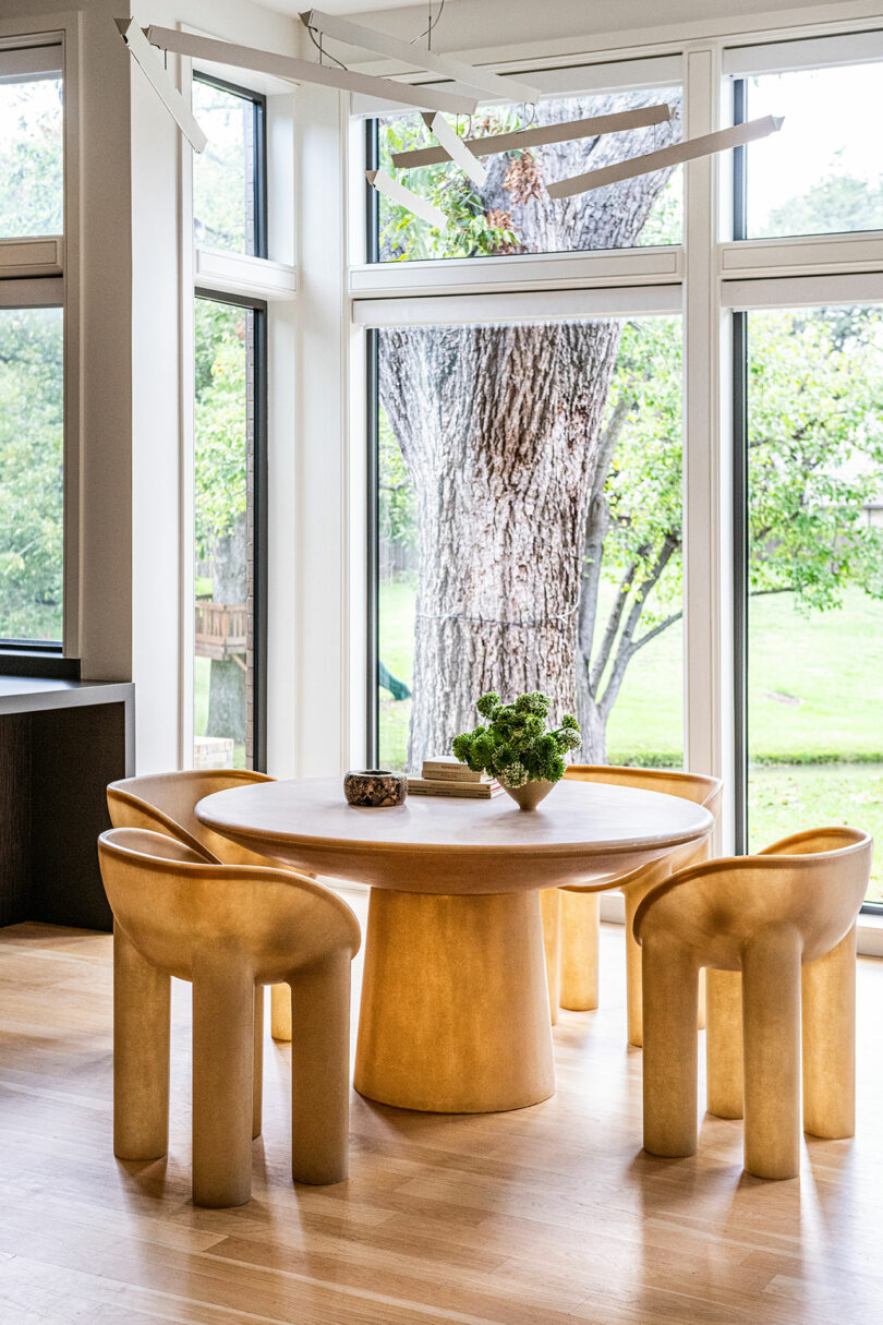 A round wooden dining table with a plant centerpiece is surrounded by four matching sculptural chairs, exemplifying Brazilian Modernism. Large windows reveal a view of a tree and green outdoor space.