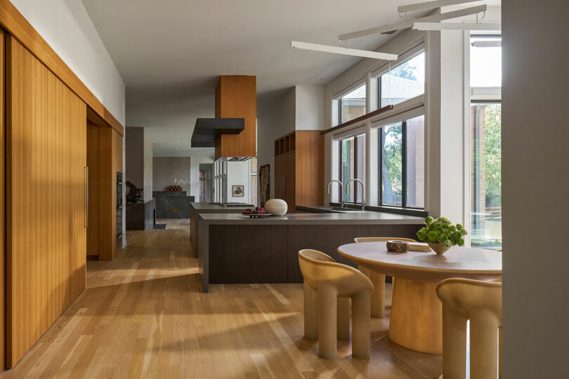 Modern kitchen and dining area with wooden flooring, wooden cabinetry, large windows, and contemporary furniture. The dining table has two beige, sculptural chairs and a green plant centerpiece, reflecting the elegance of Brazilian Modernism.