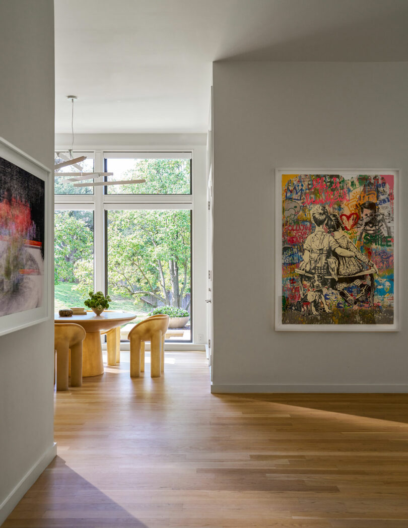 A brightly lit room with a large window, wooden floor, a dining table set with chairs, and colorful artwork and paintings on the walls exuding Brazilian Modernism.