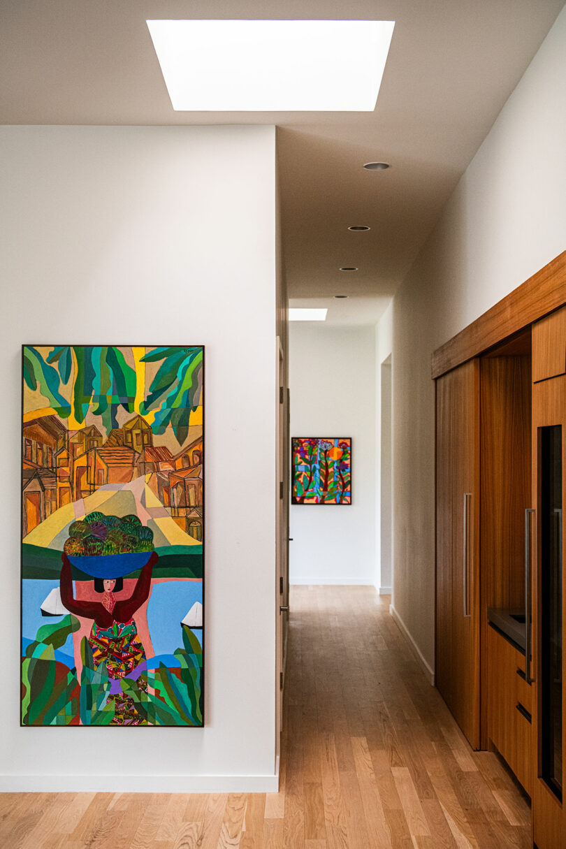 A hallway featuring colorful abstract paintings on white walls, wooden flooring, and recessed lighting exudes Brazilian Modernism. A skylight illuminates the area, complemented by sleek wooden cabinets on the right side.