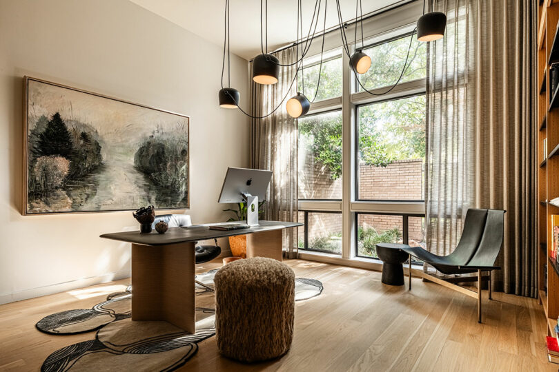 A modern home office inspired by Brazilian Modernism features a large window, abundant natural light, a desk with a computer, a painting on the wall, a pouf, a chair, and ceiling pendant lights.