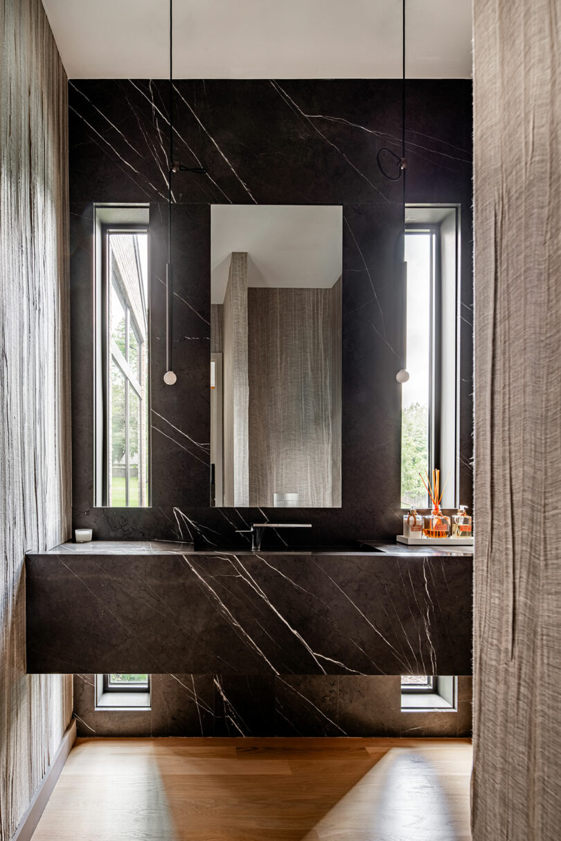 Modern bathroom with dark marble walls, rectangular mirror, black vanity, and two narrow windows on each side. Light wood flooring contrasts with the dark color scheme. Minimalist lighting fixtures hang above, embodying an essence of Brazilian Modernism in its sleek design elements.