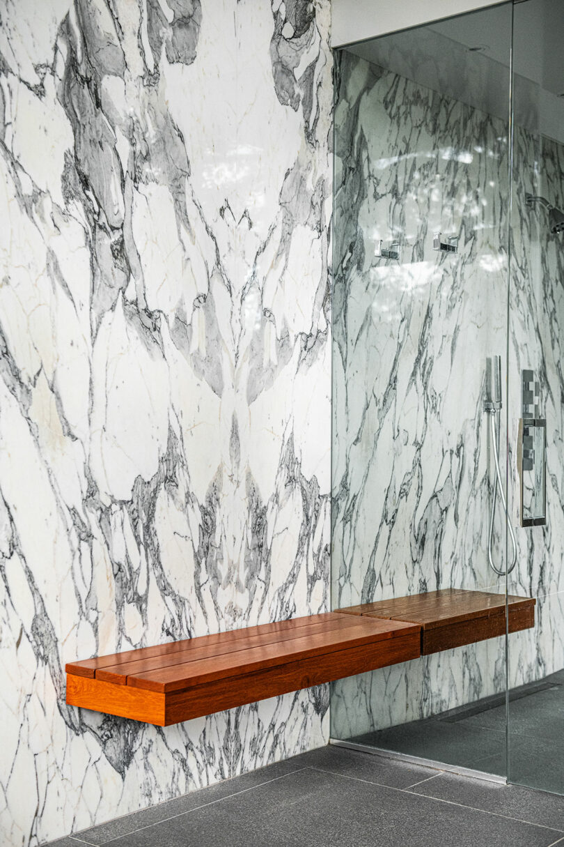 A modern shower inspired by Brazilian Modernism, featuring a large marble wall with graceful gray veining patterns, a sleek glass door, and a floating wooden bench.