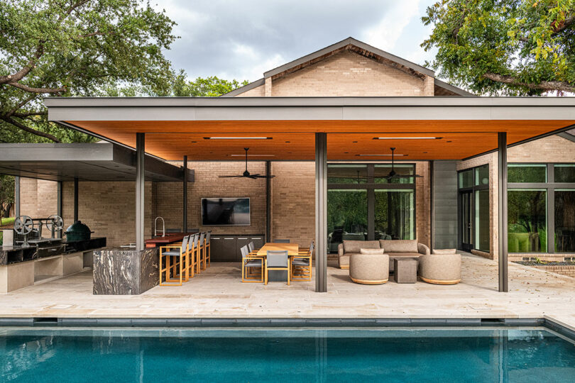 Image of a modern outdoor patio with a seating area, dining table, bar, and kitchen. Reflecting Brazilian Modernism, the space overlooks a pool and is covered by a wooden ceiling. In the background is a brick house with large windows.