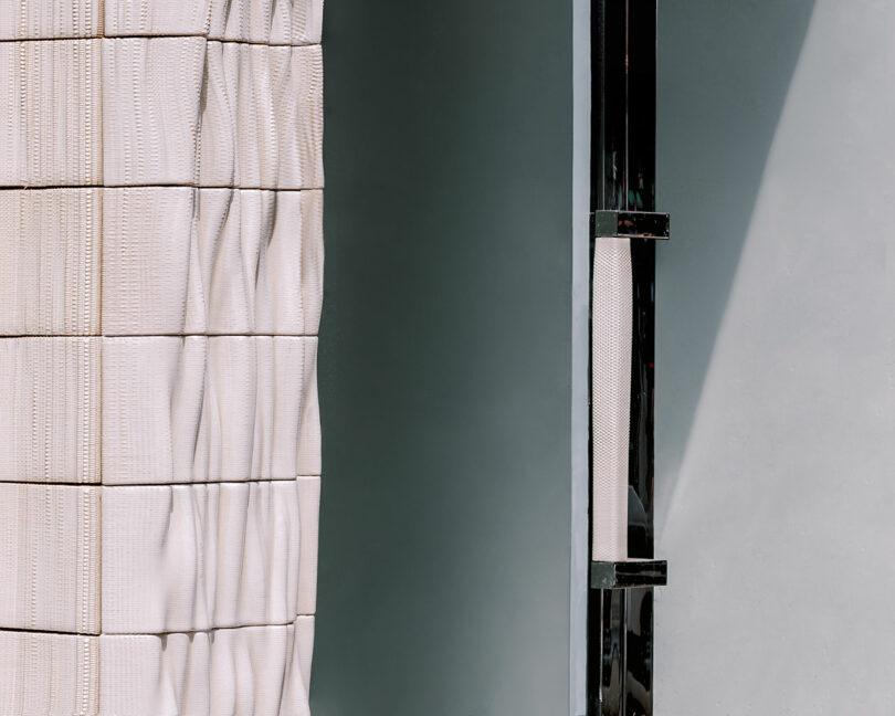 Υφασμάτινο πάνελ με υφή δίπλα σε ένα στενό κατακόρυφο γυάλινο παράθυρο σε ένα κτίριο, κάτω από έναν καθαρό, ηλιόλουστο ουρανό.