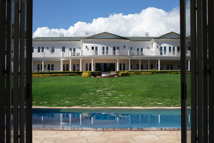 Elegant two-story white house with Design Miami symmetrical design, expansive lawn, and a foreground pool, viewed through an open gate under a blue sky with fluffy clouds.