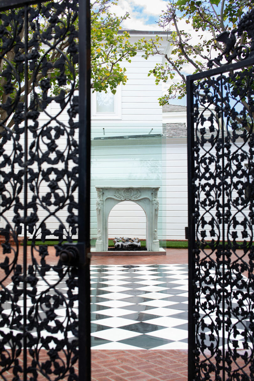 View through an ornate black iron gate focusing on a white house with a checkerboard floor patio and a small arched structure in the Design Miami garden.