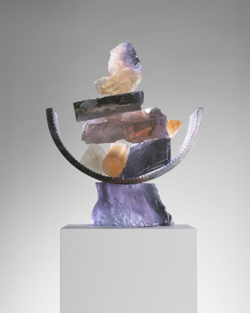 A modern sculpture featuring an arrangement of various textured rocks and minerals balanced on a curved metal base, displayed on a white pedestal against a light background at Design Miami.