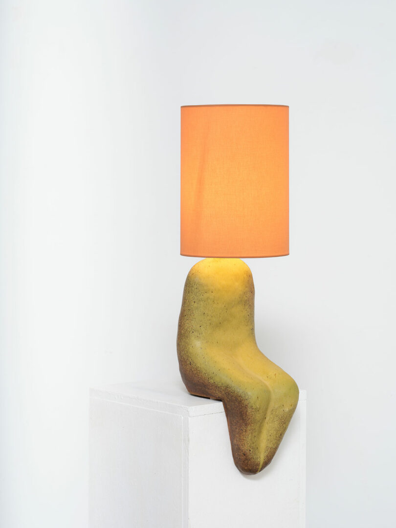 A modern lamp with an orange shade mounted on an abstract, curved, yellow and brown base, displayed at Design Miami against a white backdrop.