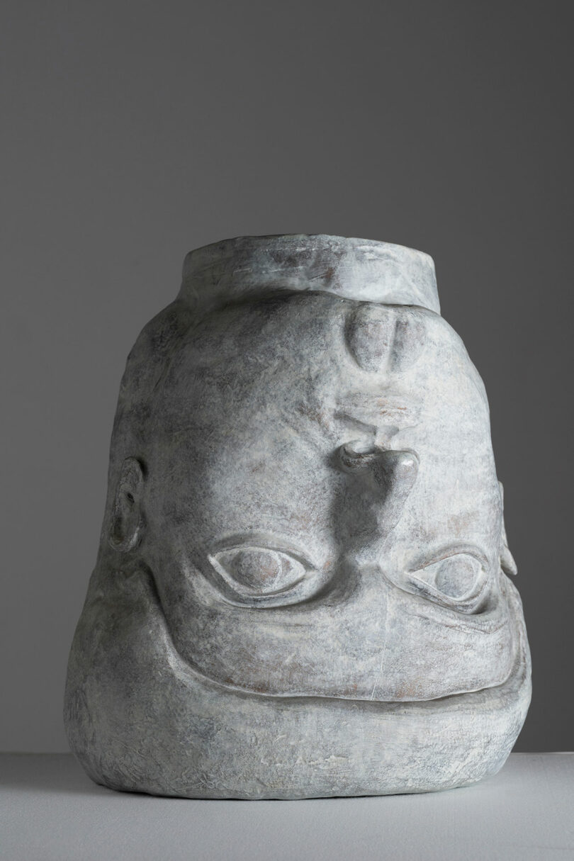 A carved stone sculpture resembling a stylized human face with large eyes and a prominent nose, featured at Design Miami, set against a gray background.