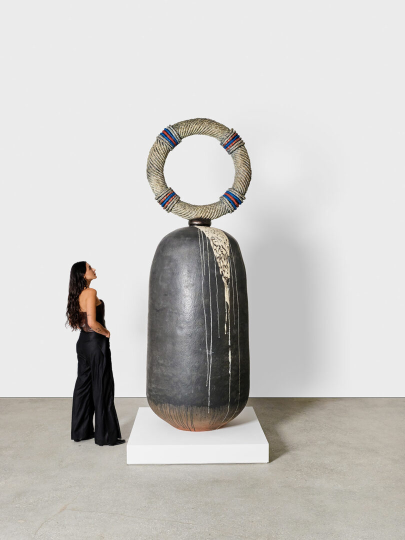 A woman stands beside a large, abstract sculpture at Design Miami, featuring a textured, cylindrical base with a ring adorned with colorful threads on top.