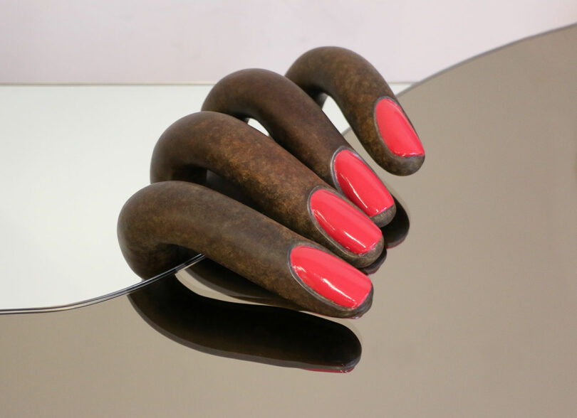 Sculpture presented at Design Miami, featuring a large metallic hand with oversized, glossy red fingernails, partially reflecting on a white surface.