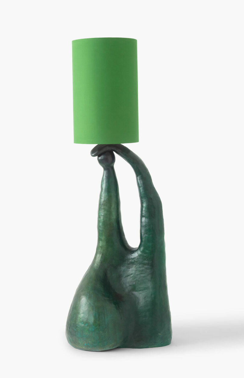 A sculpture of a hand with exaggerated fingers forming the base of a lamp, designed for Design Miami, supporting a simple green lampshade, isolated on a white background.