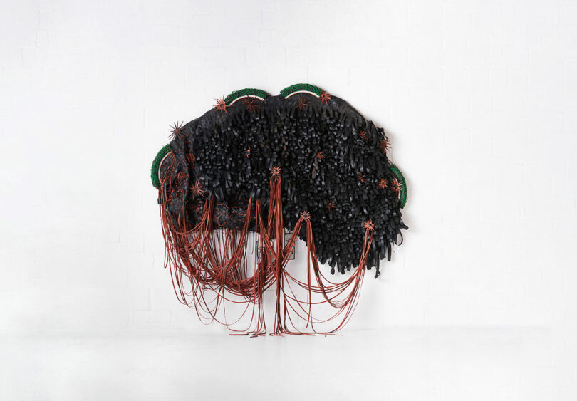Abstract wall-mounted art piece featured at Design Miami, incorporating black strands and red cables cascading from multiple circular green bases on a white brick wall.