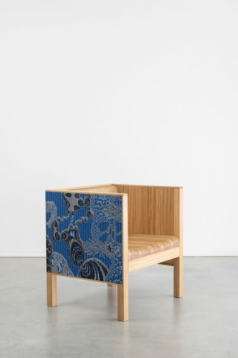 A modern wooden armchair with a blue patterned upholstery on one side, positioned on a plain white background at Design Miami.
