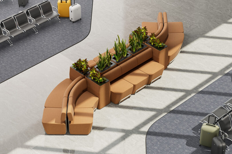 Aerial view of an airport lounge with curved orange modular seating with green plants. The area is surrounded by grey flooring and scattered black and white suitcases.