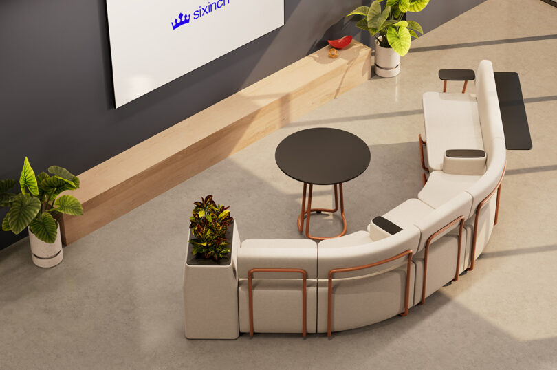 Modern office lounge area with a modular curved white couch, round black table, and plants. The wall features a screen with the text "sixinch." Light wood accents and a large potted plant are also present.