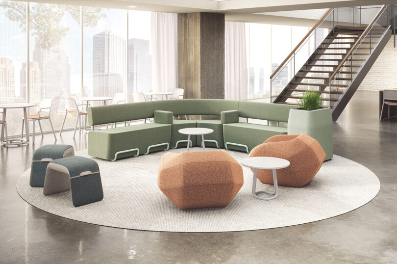 Modern office lounge with curved green modular sofa, orange and gray ottomans, small round tables, and a staircase. Floor-to-ceiling windows reveal cityscape in the background.