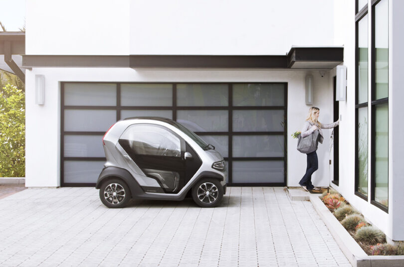 A woman with a grocery bag opening the garage door to park a micro EV, Eli ZERO, in a modern home driveway.