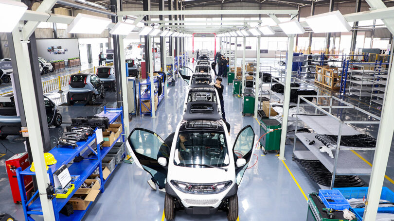 Interior view of a car manufacturing plant with multiple Eli ZERO micro EVs in assembly lined up, equipment, and workers visible.