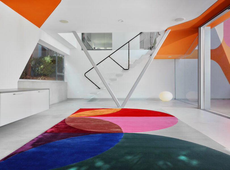 Modern interior with a colorful circular carpet, white walls, an orange curved ceiling, and a staircase with black railings