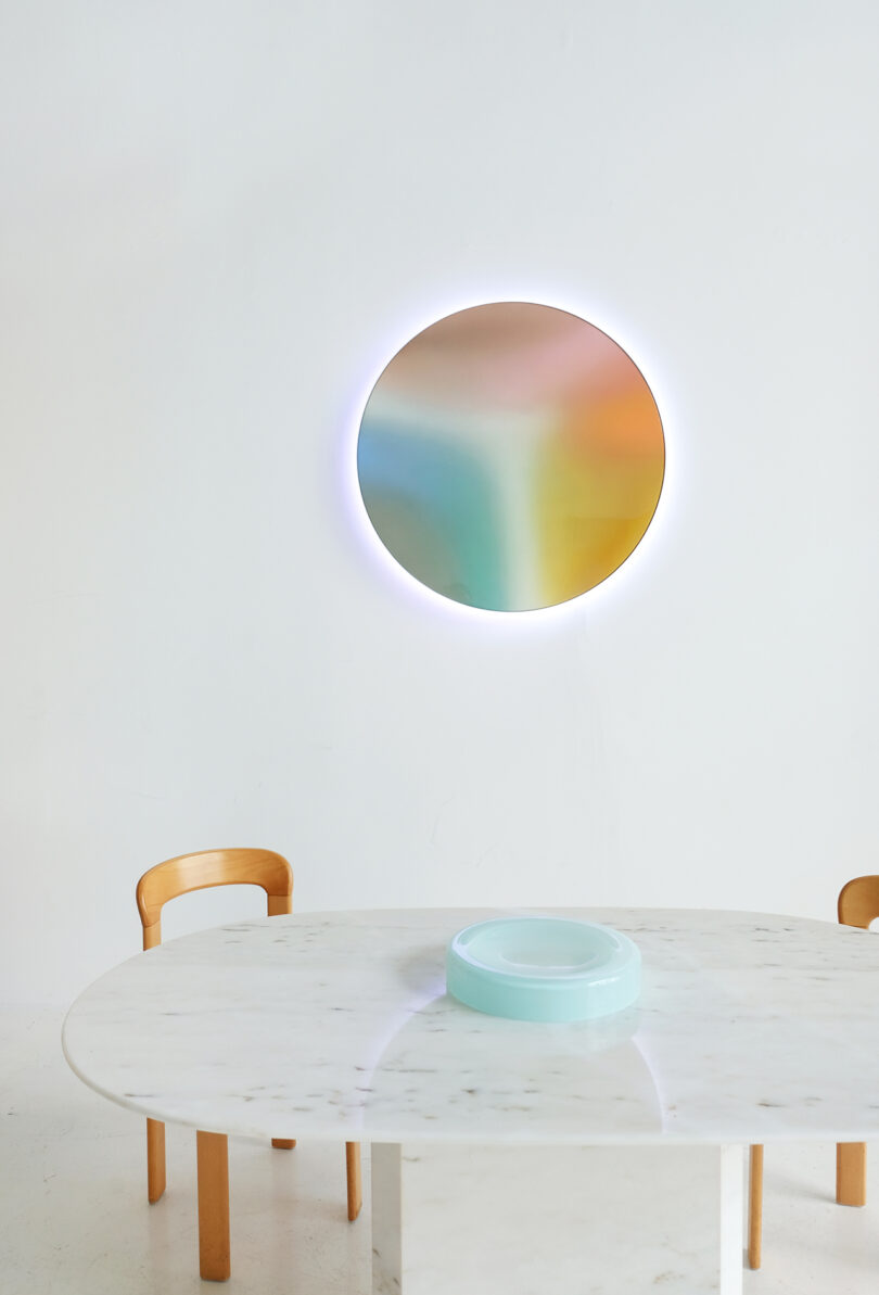 A modern dining room featuring a marble table with curved wooden chairs and a colorful circular light fixture on a white wall.