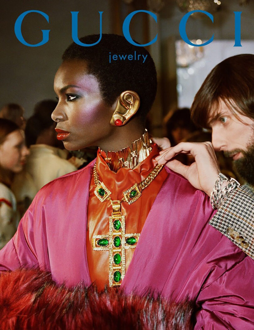 A model poses for a jewelry advertisement, wearing a colorful silk outfit and elaborate earrings, with a male figure adjusting her necklace in the background.