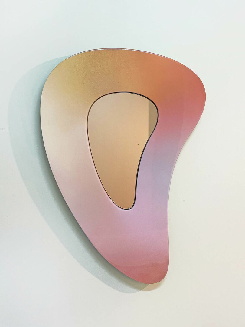 Modern abstract wall mirror featuring a layered, teardrop-shaped piece with a gradient from peach to pink tones.