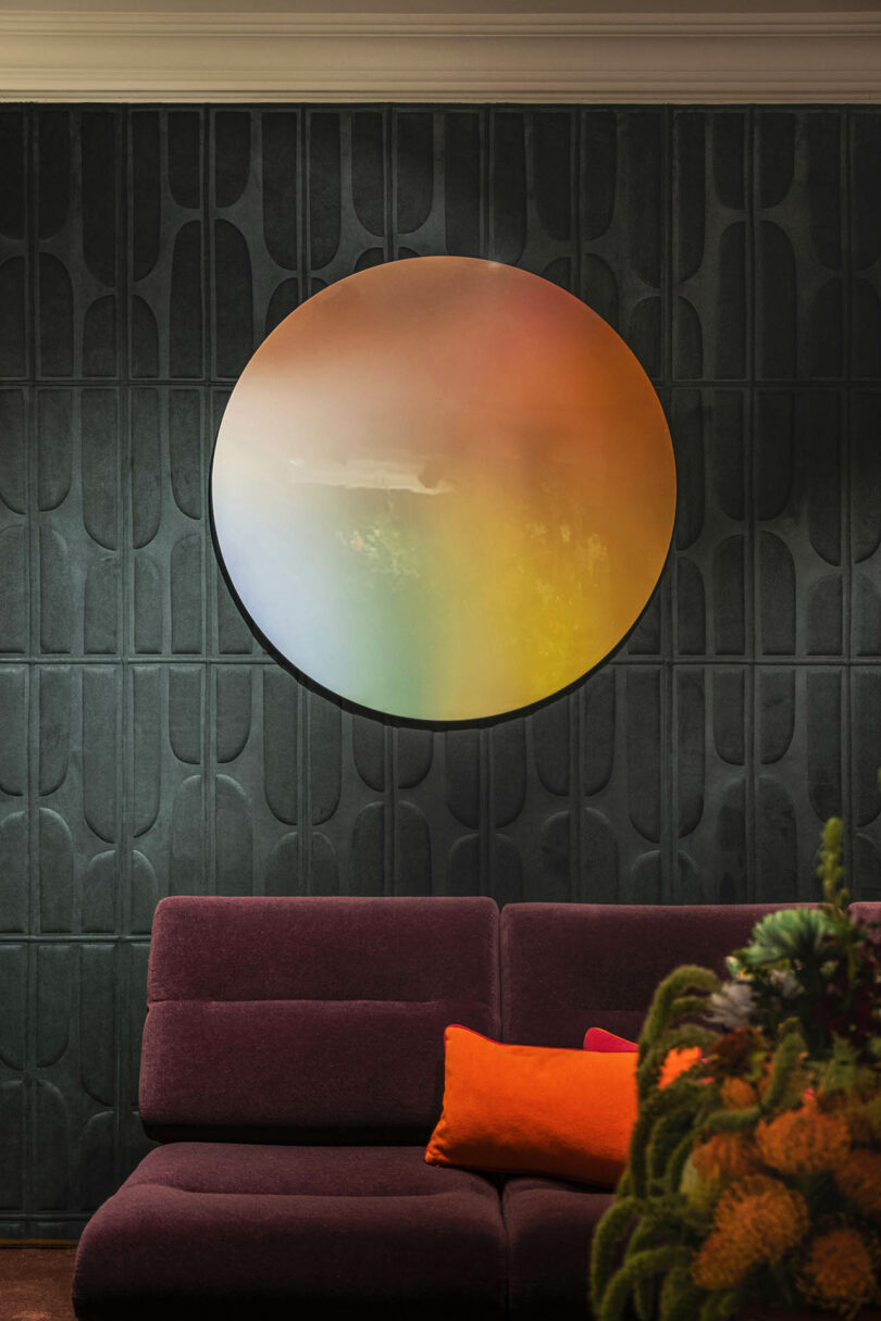 A large, circular, multicolored abstract piece of art hangs above a deep purple sofa with an orange cushion in a room with dark paneled walls.