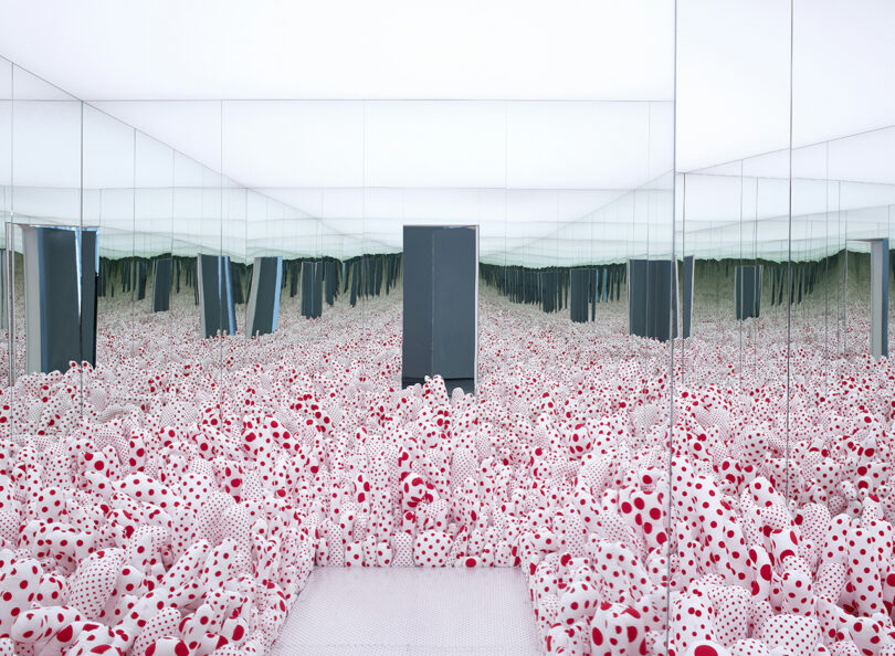 An art installation featuring a mirrored room filled with numerous red spheres with white polka dots, punctuated by black columns.