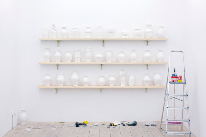 A white wall displays shelves of white ceramic vases. A stepladder stands to the right, with tools and equipment on the floor below.