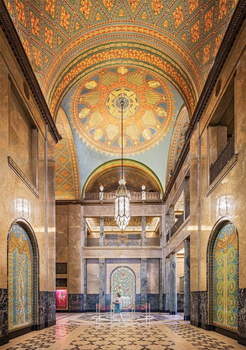 A grand lobby with an ornate, colorful domed ceiling, marble floors, arched doorways, and a large chandelier. A blurred figure is walking in the center of the space.