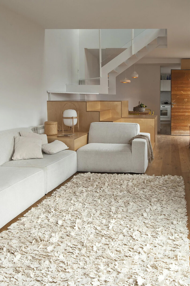 A modern living room with a white sectional sofa, a textured white rug, and a wooden staircase leading to an upper level. The space also includes a wooden dining area and a partially visible kitchen.