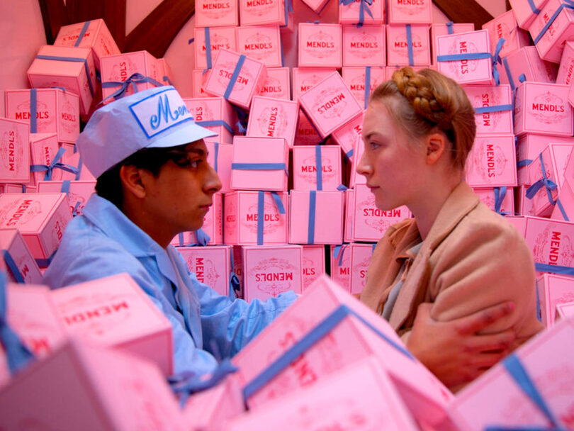 Two people converse surrounded by a large pile of pink boxes with blue ribbons labeled "Mendl's". One wears a blue uniform and hat, and the other has a braided hairstyle and beige coat.