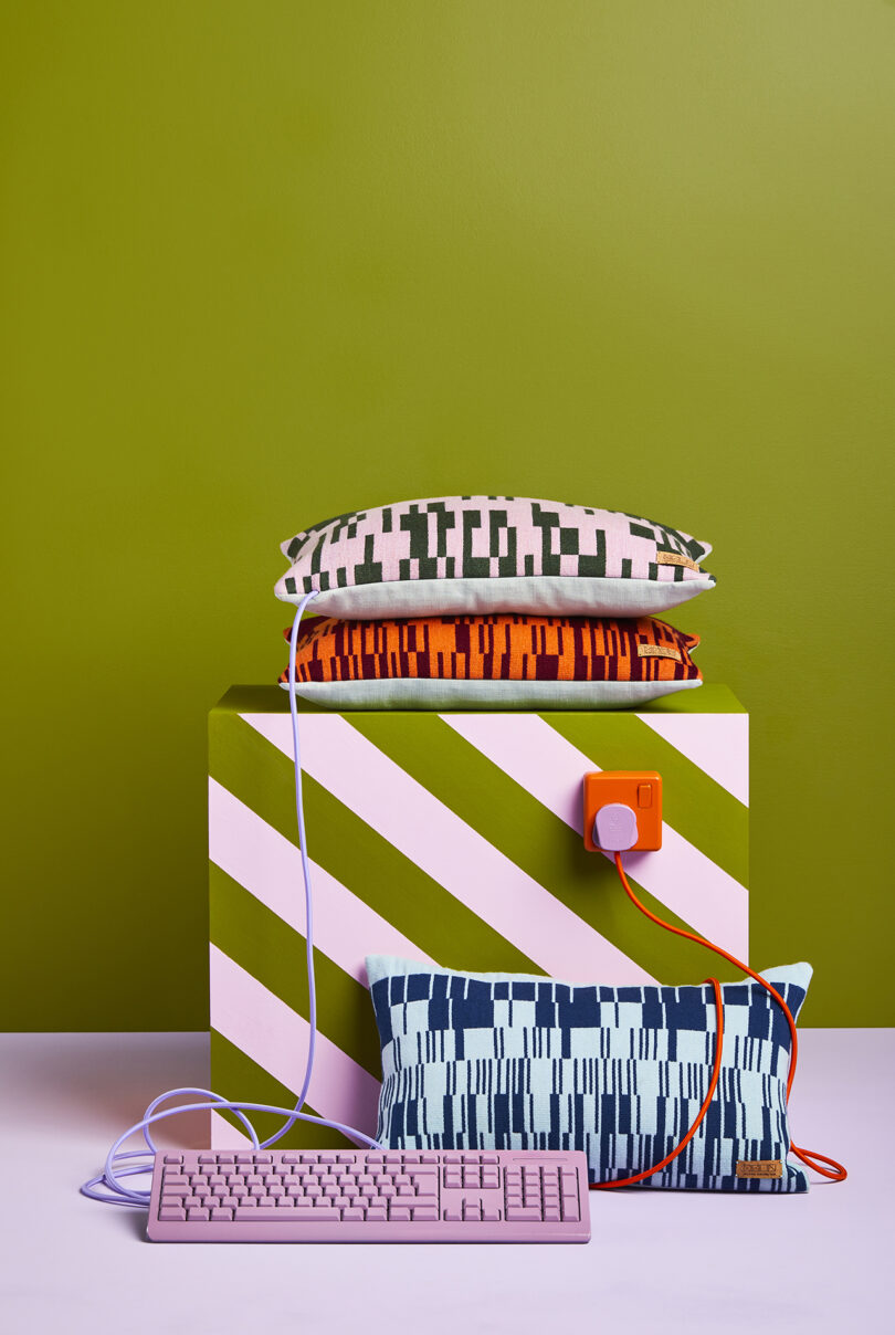 A stack of patterned pillows rests on a pink and green striped box, with a blush pink keyboard and orange power brick connected by lavender and orange cables in front.