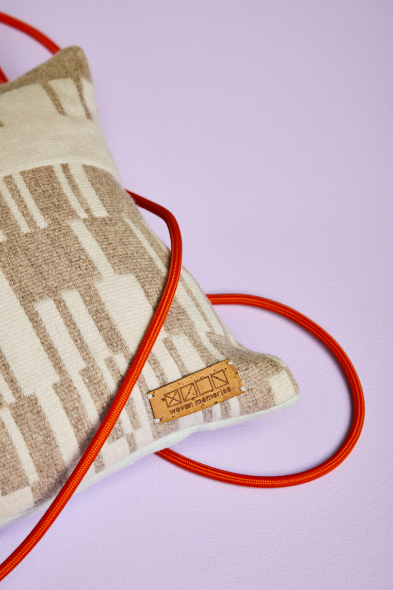 Close-up of a beige patterned cushion with a brown leather tag and a red cord on a light purple surface.