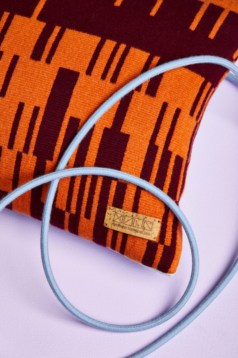 Close-up of a cushion with an orange and dark red pattern and a light blue cord placed on top. A small tag with decorative text is sewn onto the cushion. The background is light purple.