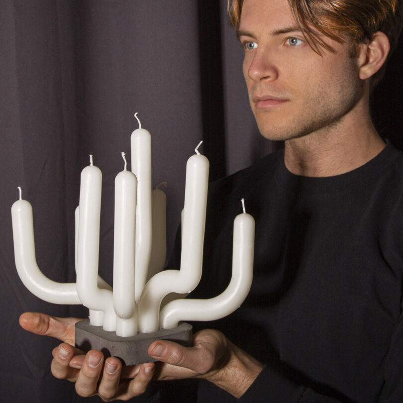 Man holding a candelabra with seven unlit white candles, wearing a black shirt against a dark background.