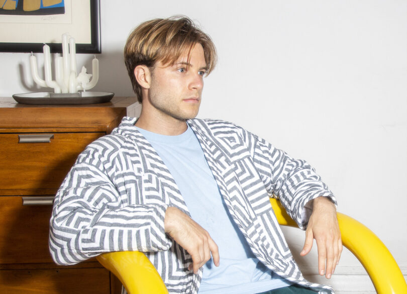 Man with a bowl haircut sitting on a yellow chair, wearing a blue t-shirt and a patterned button-down, looking to the side in a white room.
