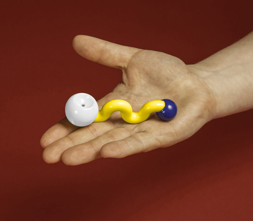 A hand holding a colorful, twisted pipe with white, yellow, and blue sections, set against a red background.