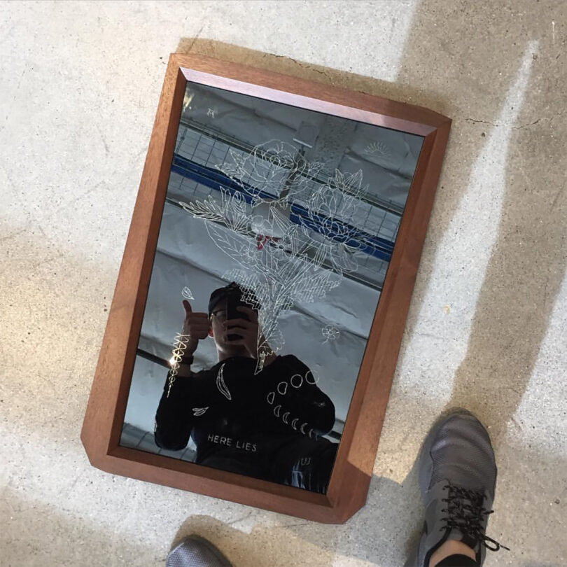 A person in dark clothing gives a thumbs-up reflected in a rectangular framed mirror with floral engravings, placed on a concrete surface next to a pair of feet in gray shoes.