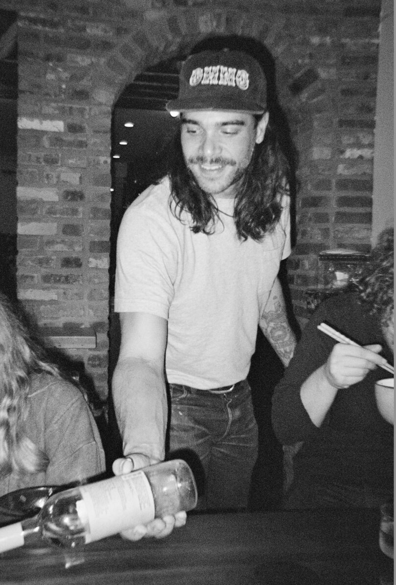 Man wearing a cap and a T-shirt holding an empty wine bottle at a dining table with people eating. Brick wall in the background. Black and white photo.