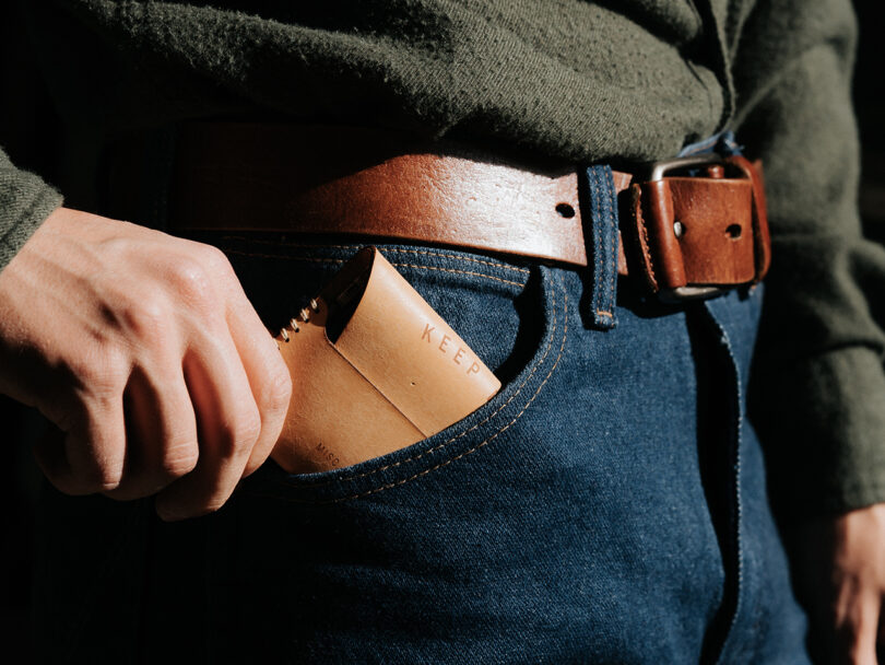A person wearing a green shirt and blue jeans with a brown leather belt is placing a tan leather wallet into their front pocket.