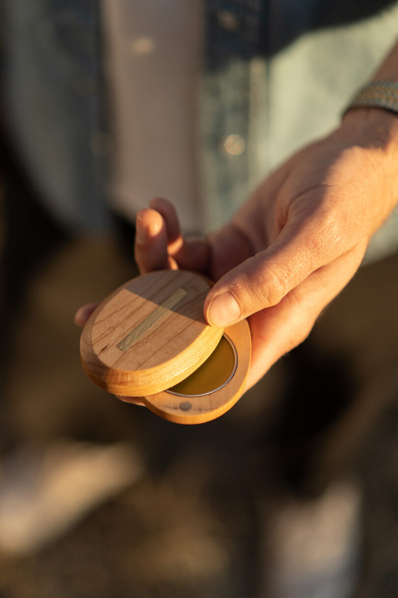 A person holding a round, wooden container with a hinged lid, revealing solid perfume.
