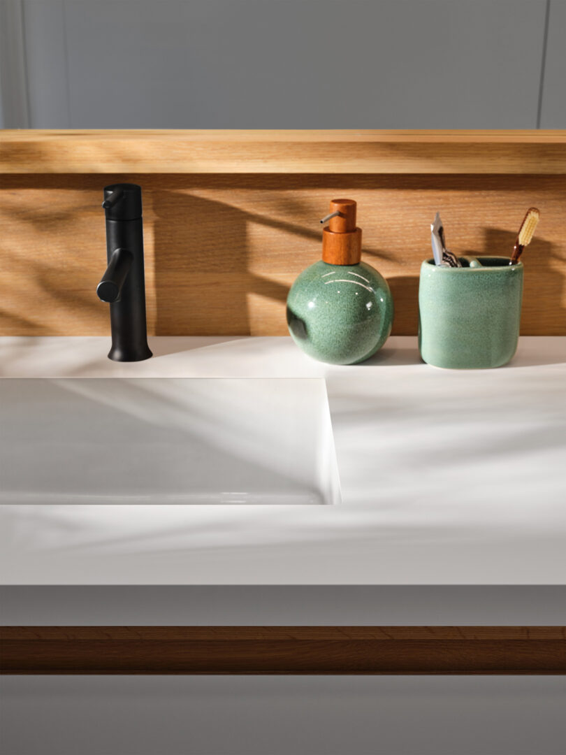 A bathroom countertop with a white sink, black faucet, a green soap dispenser, and a green container holding toothbrushes, set against a wooden backsplash.
