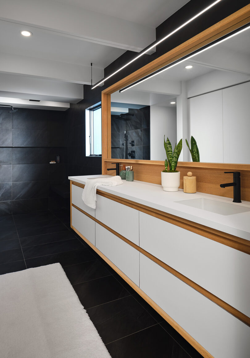 Modern bathroom with white vanity, large mirror with wooden frame, potted plant, and black fixtures. White towel on counter; black and white tiles on floor and wall; illuminated by overhead lights.