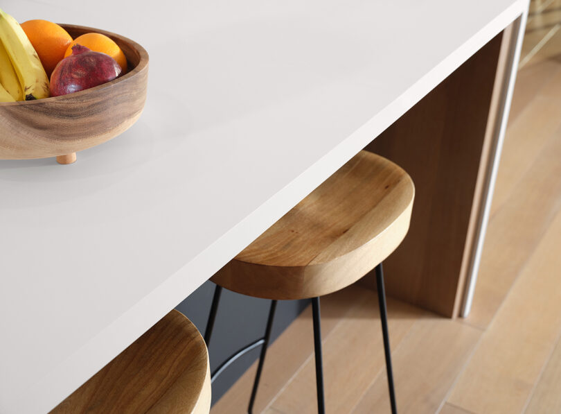 A wooden fruit bowl containing bananas, an apple, and an orange sits on a white countertop next to two wooden bar stools.