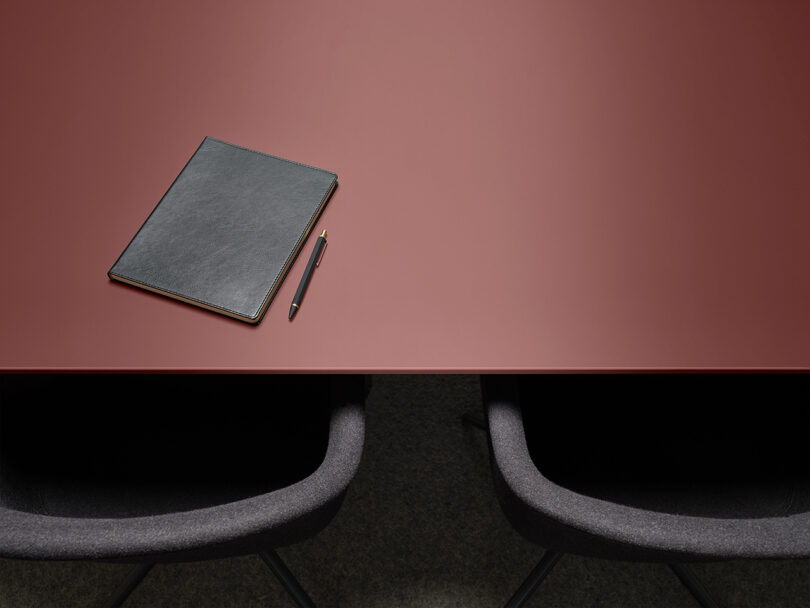 A dark red table with two grey chairs. On the table, there is a closed black leather notebook and a black pen.