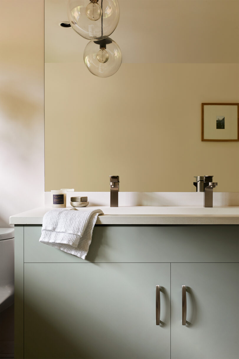 Modern bathroom with a large mirror, a light green vanity with metal handles, a white countertop with folded towels and toiletries, and a wall-mounted light fixture with round glass shades.